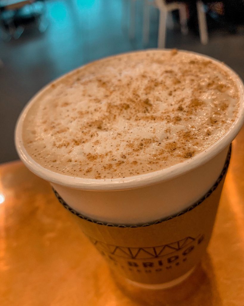 Latte in a white paper cup with a brown Blue Bridge Coffeehouse-branded sleeve on a wooden table