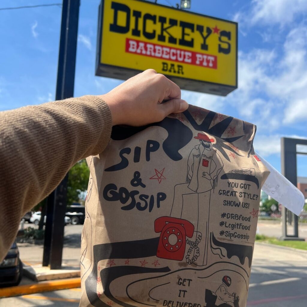 Dickey's BBQ to-go order bag being held up in front of the Dickey's sign with blue sky in the background