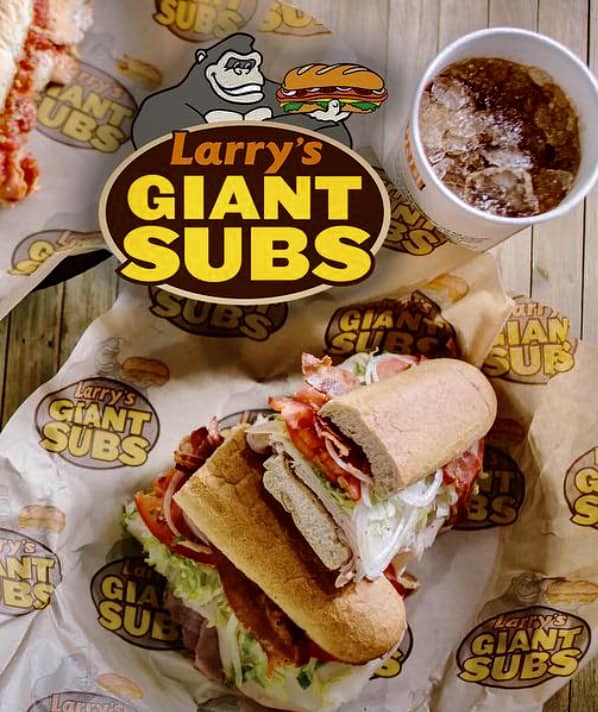 Larry's Giant Subs Logo on a photo of a Bacon Turkey Sub sandwhich with lettuce tomato and onion on a whole whet roll with a cola in a cup on the side