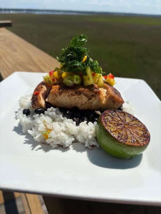 Blackend Mahi Mahi on a bed of black beans and white rice topped with pineapple mango salsa and parsley garnish and a half a lime that's been seared on the cut side--all on a white plate sitting on a wood railing with the salt marsh in the background