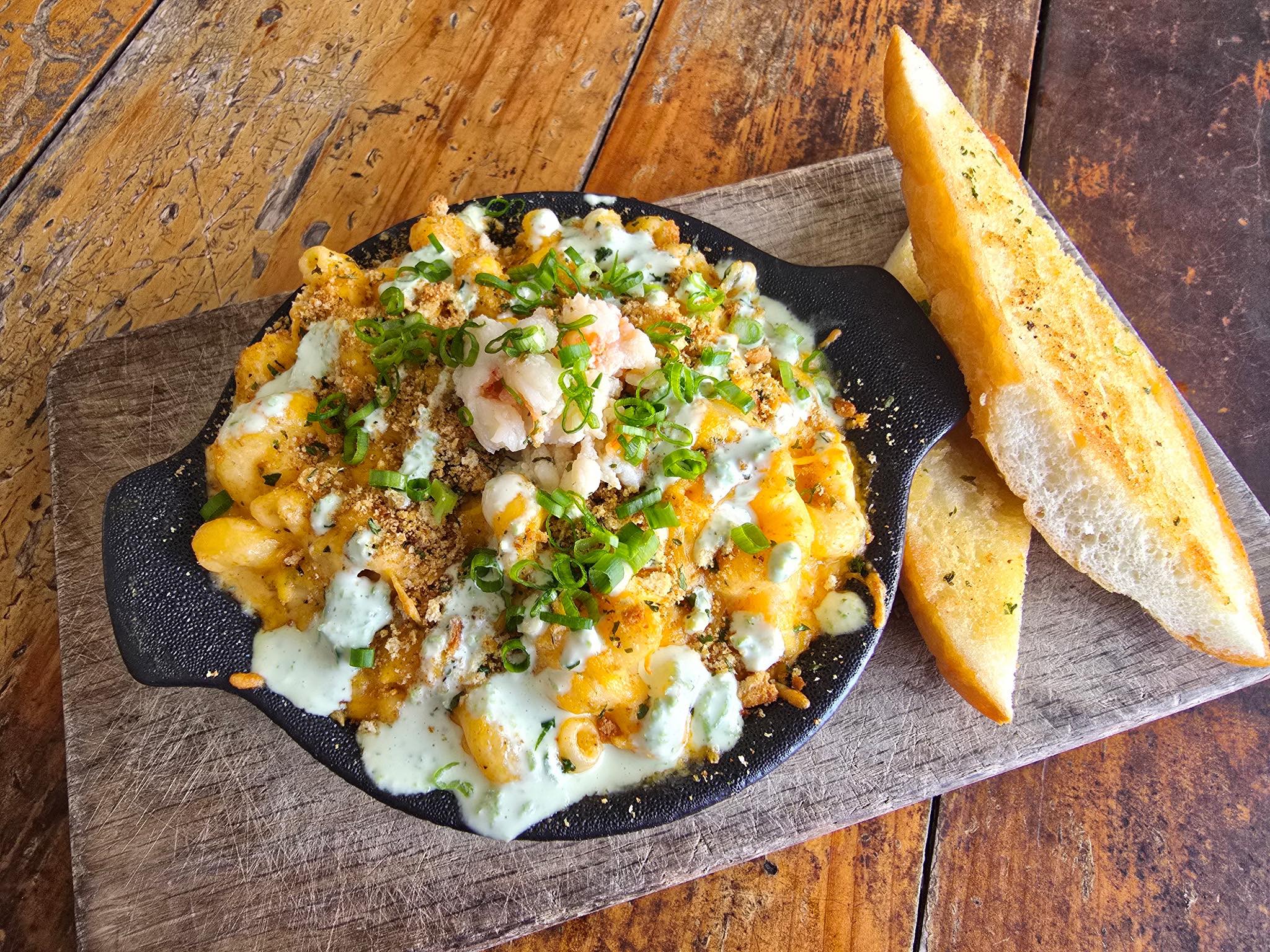 Round cast iron bowl of lobster macaroni and cheese garnished with minced green scallion, all sitting on a rood cutting board with two toasted slices of garlic bread
