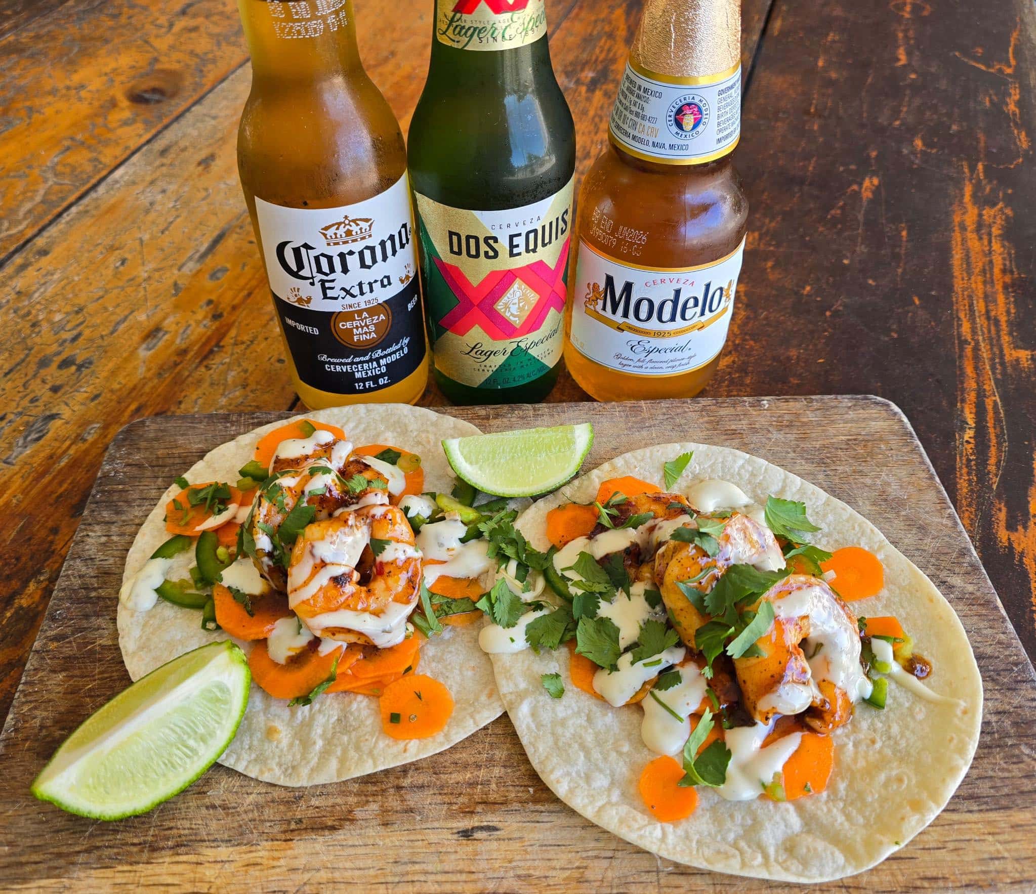 Two open faced shrimp tacos on a cutting board with cilantro and lime wedge garnishes with three brands of bottled Mexican beer (Corona Extra, Dos Equis, and Modelo) in the background