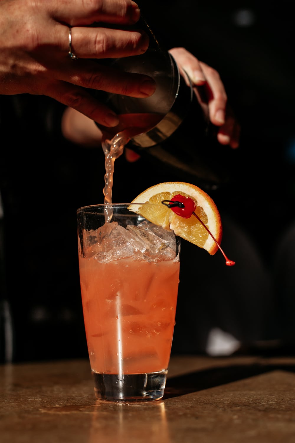 Orange colored cocktail being poured into a clear tall glass with ice and an orange slice and cherry garnish