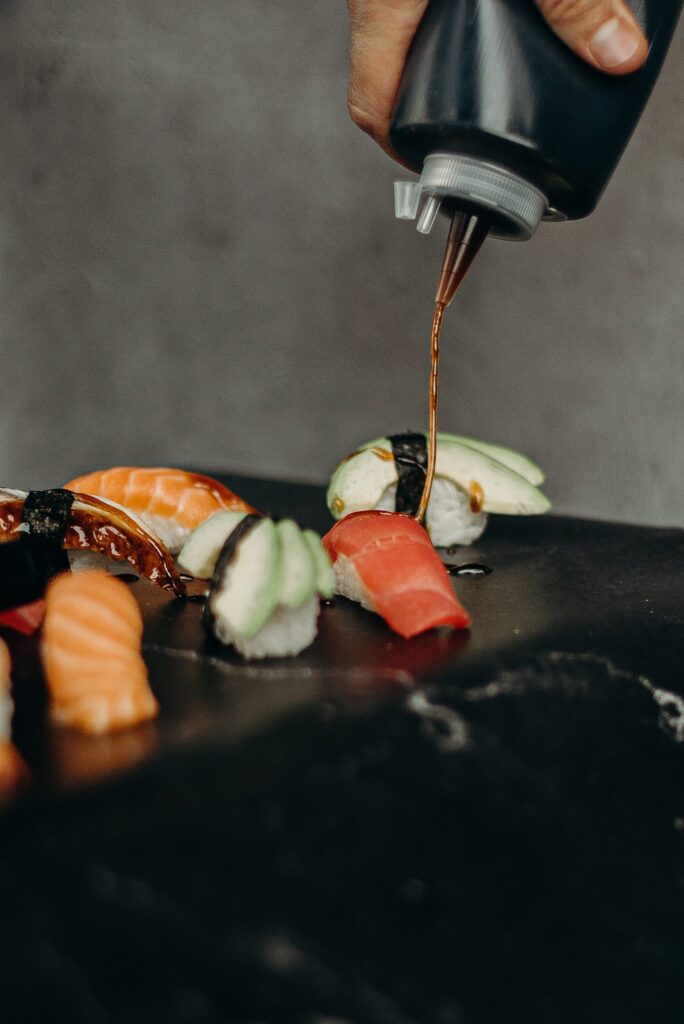 A hand squeezing a bottle of soy sauce onto five different kinds of sushi including shrimp, salmon, and avocado