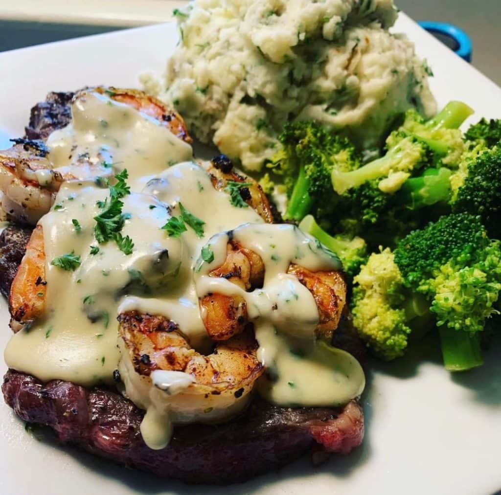 Grilled steak topped with grilled shrimp and covered with Hollandaise sauce with sides of steamed broccoli and mashed potatoes on a white plate at Outerbanks Bar and Grill