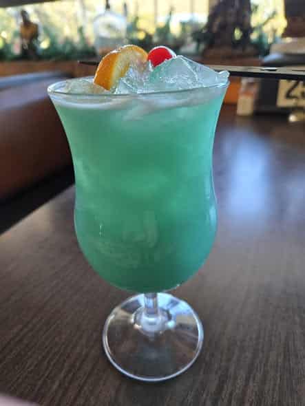 Aqua green colored cocktail named a "tipsy turtle" in a clear glass with orange wedge and cherry garnish