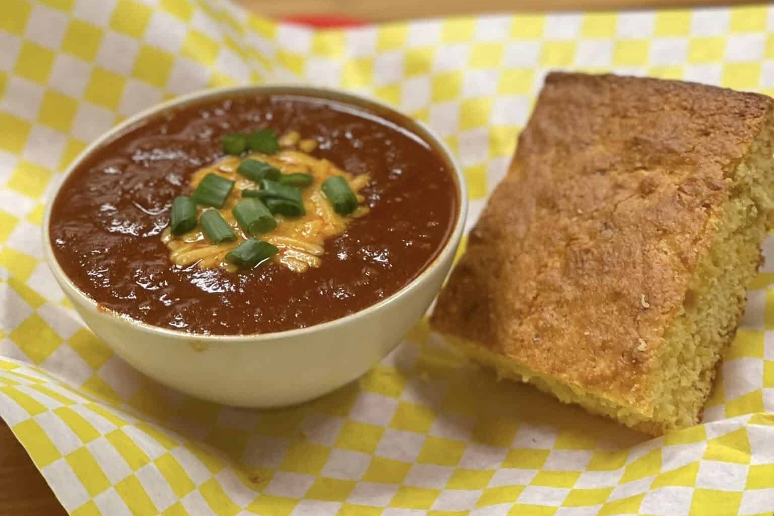 White cup of chili topped with shredded cheddar cheese and chopped scallions with a side of cornbread sitting on white and yellow checkered parchment paper
