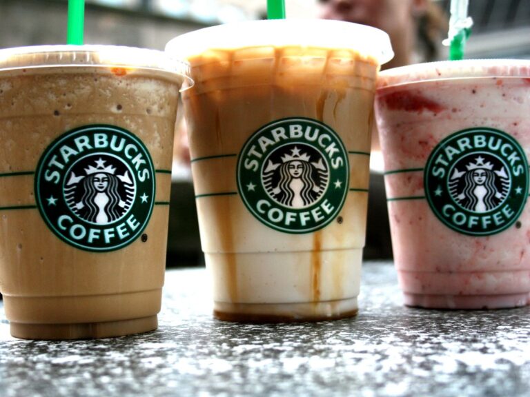 Three iced lattes in clear plastic Starbucks branded cups with lids and green straws on a granite counter top