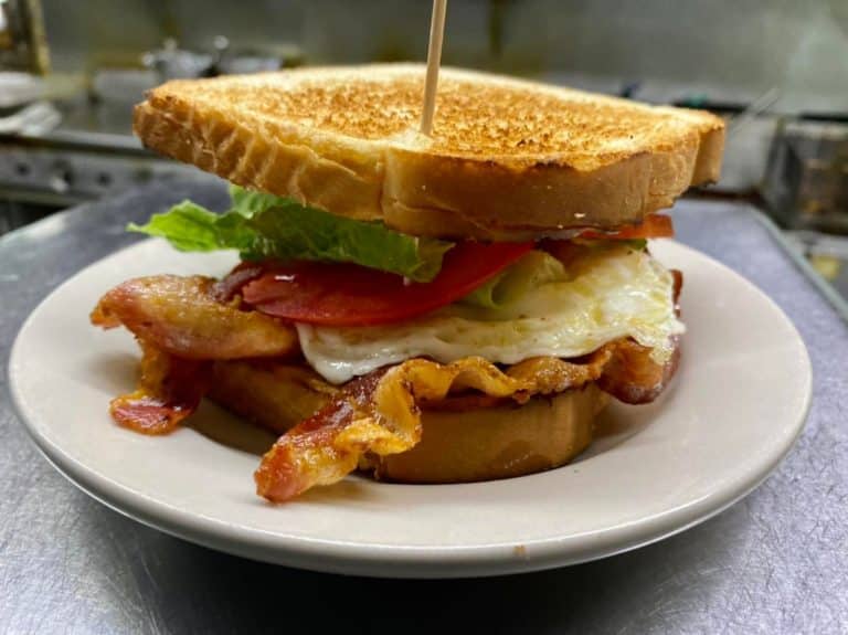 Fried egg and bacon sandwich on toasted white bread with lettuce and tomato--all pierced by a toothpick and sitting on a white plate