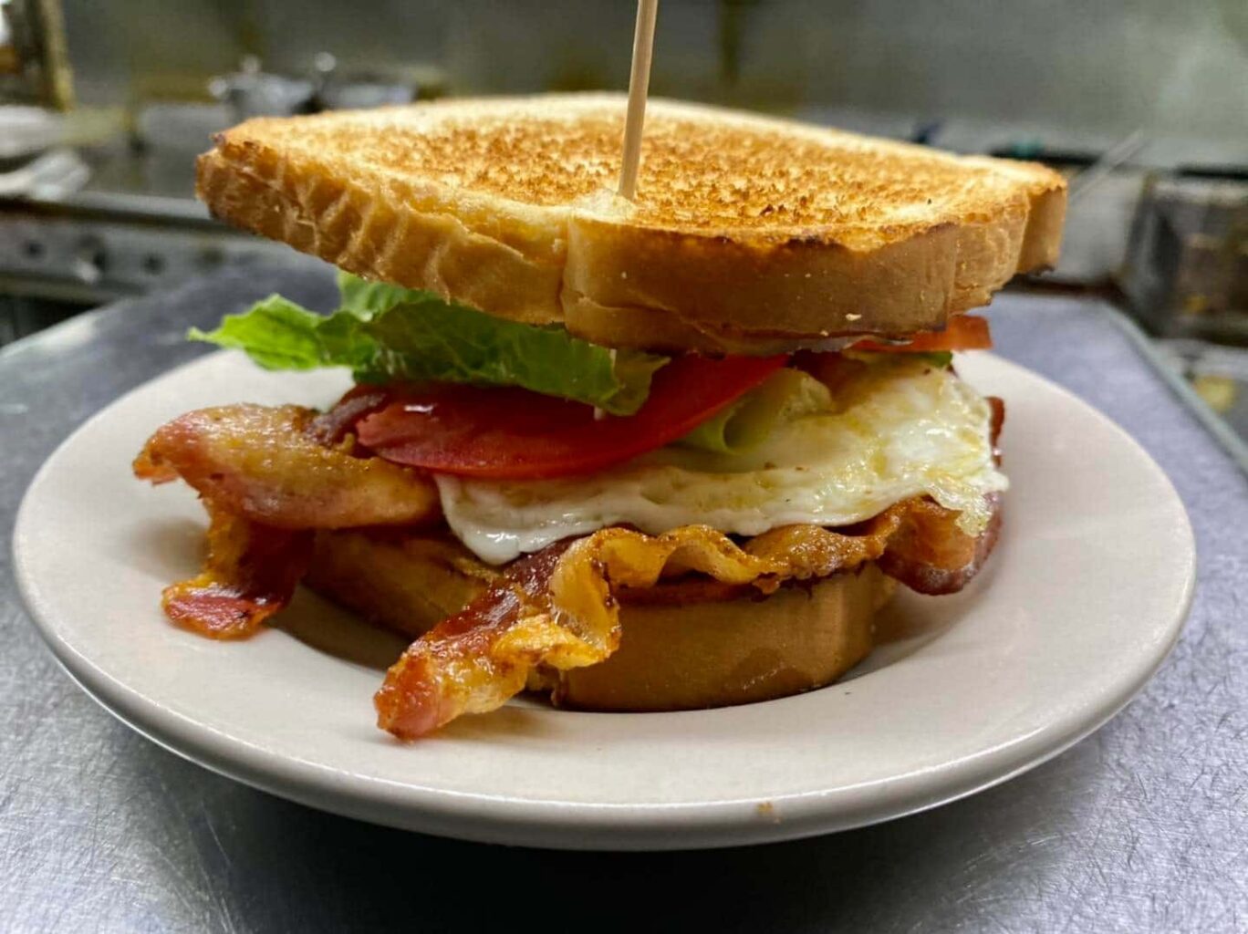Fried egg and bacon sandwich on toasted white bread with lettuce and tomato--all pierced by a toothpick and sitting on a white plate