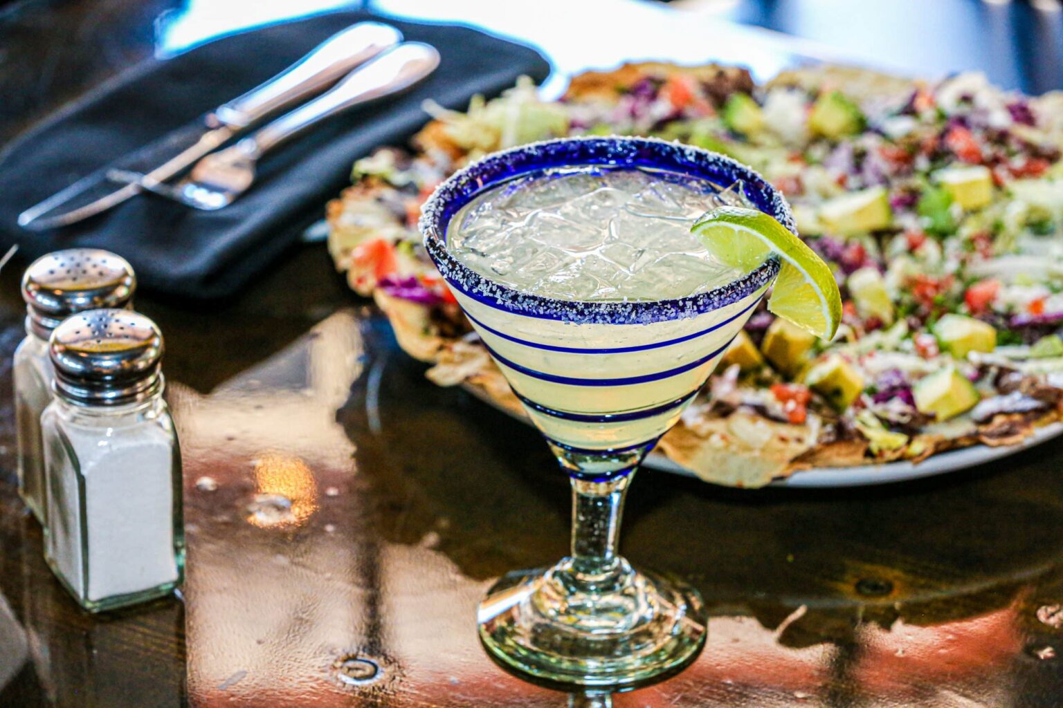 Iced Margarita with lime wedge in a hand-blown margarita glass with navy blue spiral with a plate of loaded nachos in the background and two salt shakers on the side