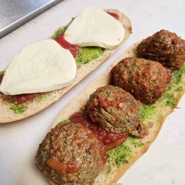 Open-face pesto meatball slider on a sub bun with pesto, Roma tomatoes, and mozzarella on one side, and pesto, four large meatballs, and marinara sauce on the other