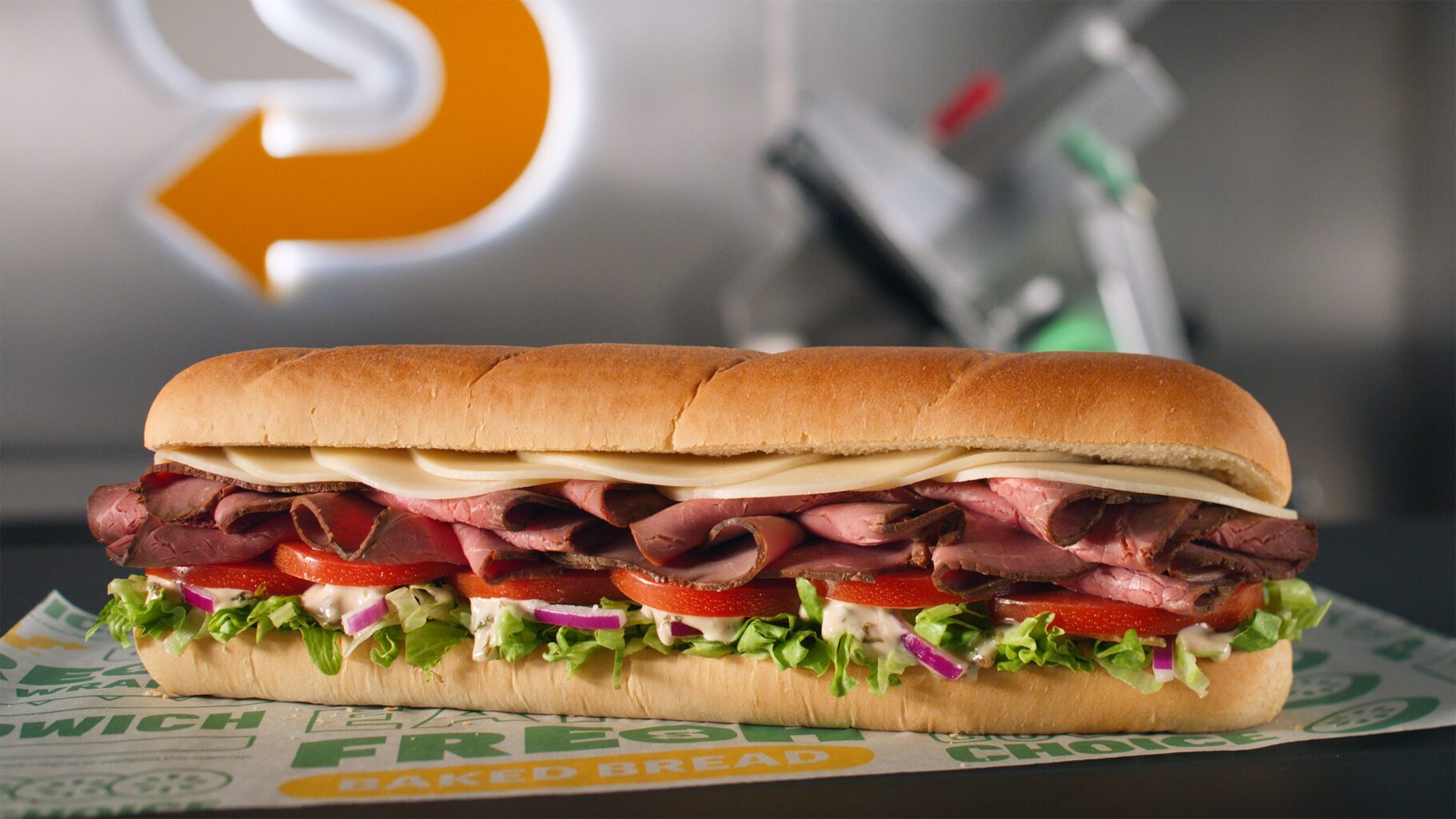 Whole Subway sandwich with roast beef, provolone cheese, lettuce and tomato on a white roll sitting on a counter