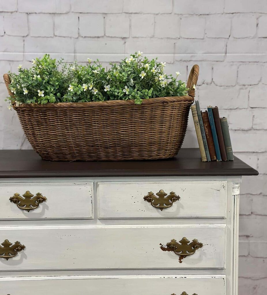 Flowering plants in a brown wicker basket on top of a white dresser with brass hardware and a black top in front a whitewashed brick wall at Cottle and Gunn. Several old books are leaning up against the basket