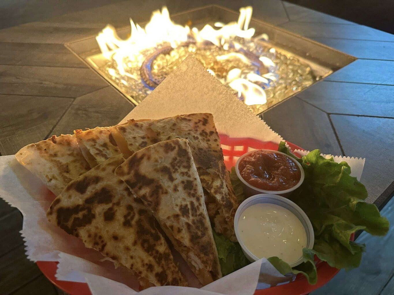 Quesadilla wedges in a red basket with sides of salsa and ranch dressing in front of a lit gas fire pit at night