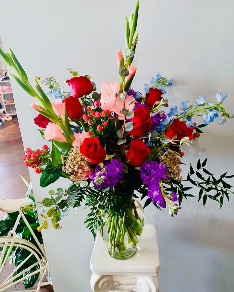 Bouquet of red roses, pink gladiolas, and other flowers and greens in a clear vase on a white plaster pedestal.