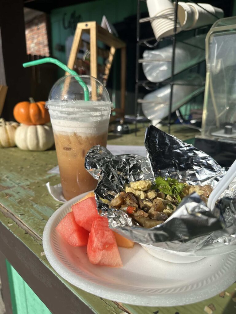 Breakfast scramble in aluminum lined basket on a white plate with chunks of watermelon on the side; also an iced coffee drink in a clear plastic cup with lid and a green straw in the background, all sitting on a counter
