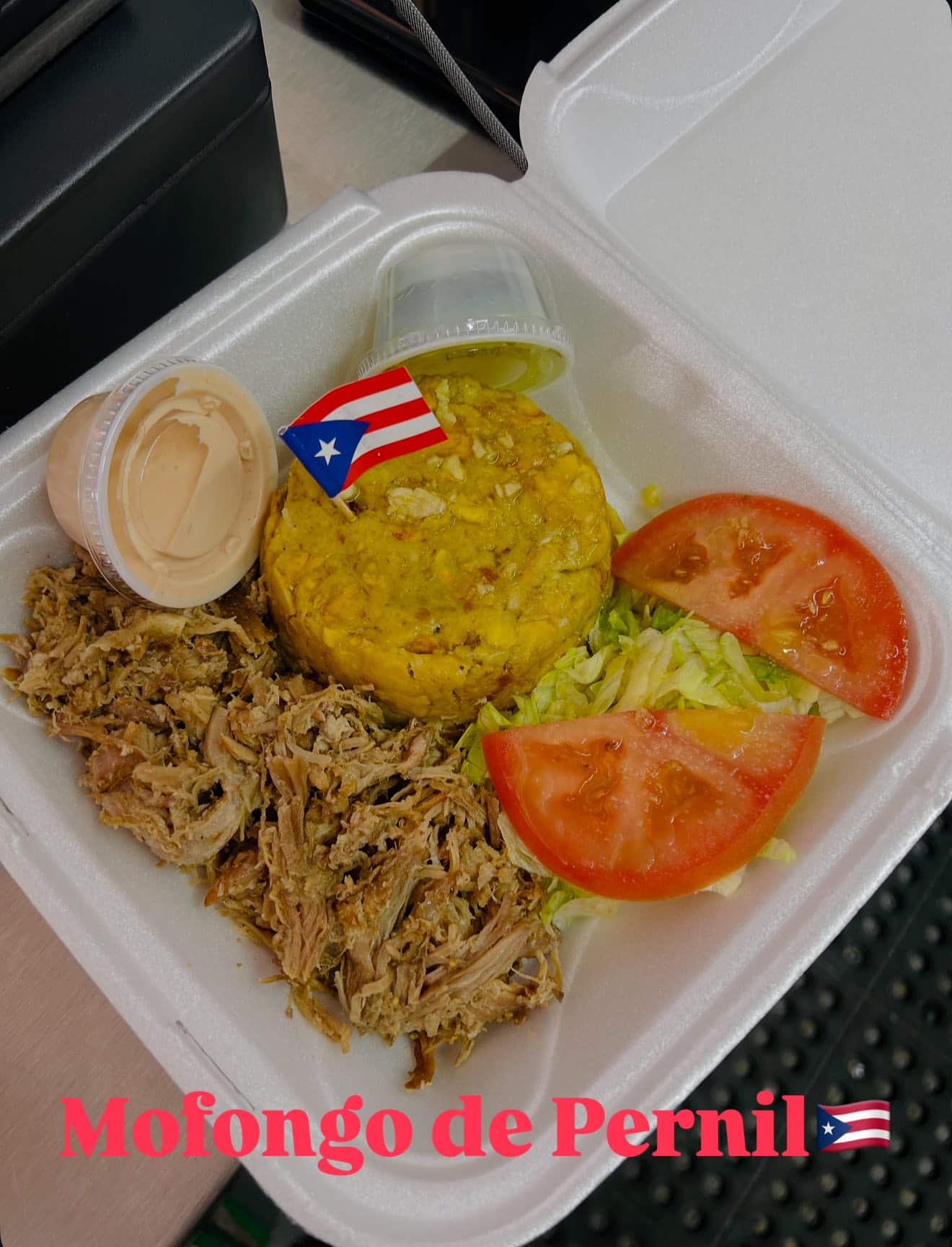 A Styrofoam container with pulled pork, mufongo with a tiny paper Puerto Rican flag on a toothpick, a side of shredded lettuce and sliced tomato, and two small plastic containers of sauces.