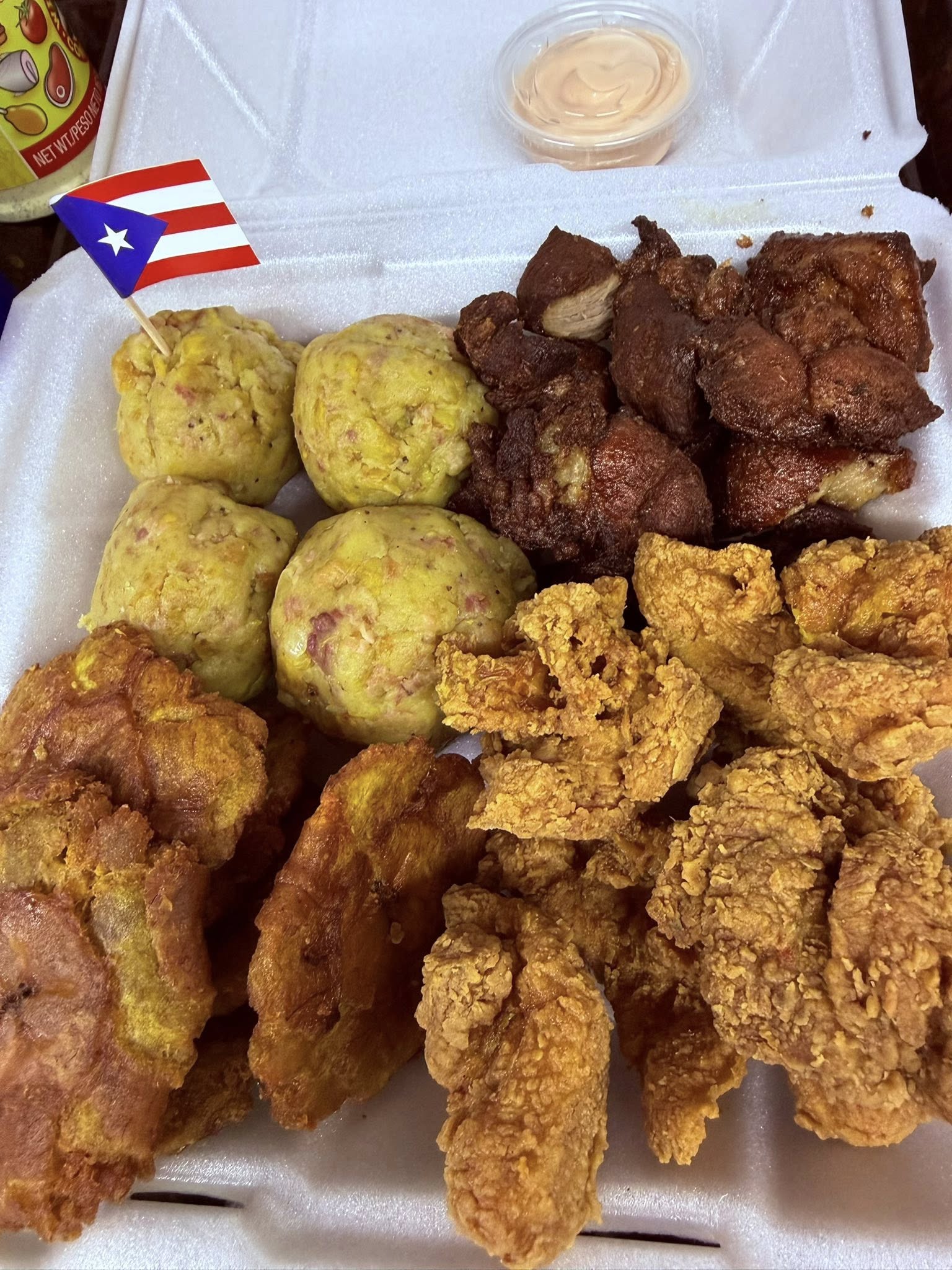 A Styrofoam box container with mufongo, fried beef tips, fried chicken strips and with a Puerto Rican flag on a toothpick