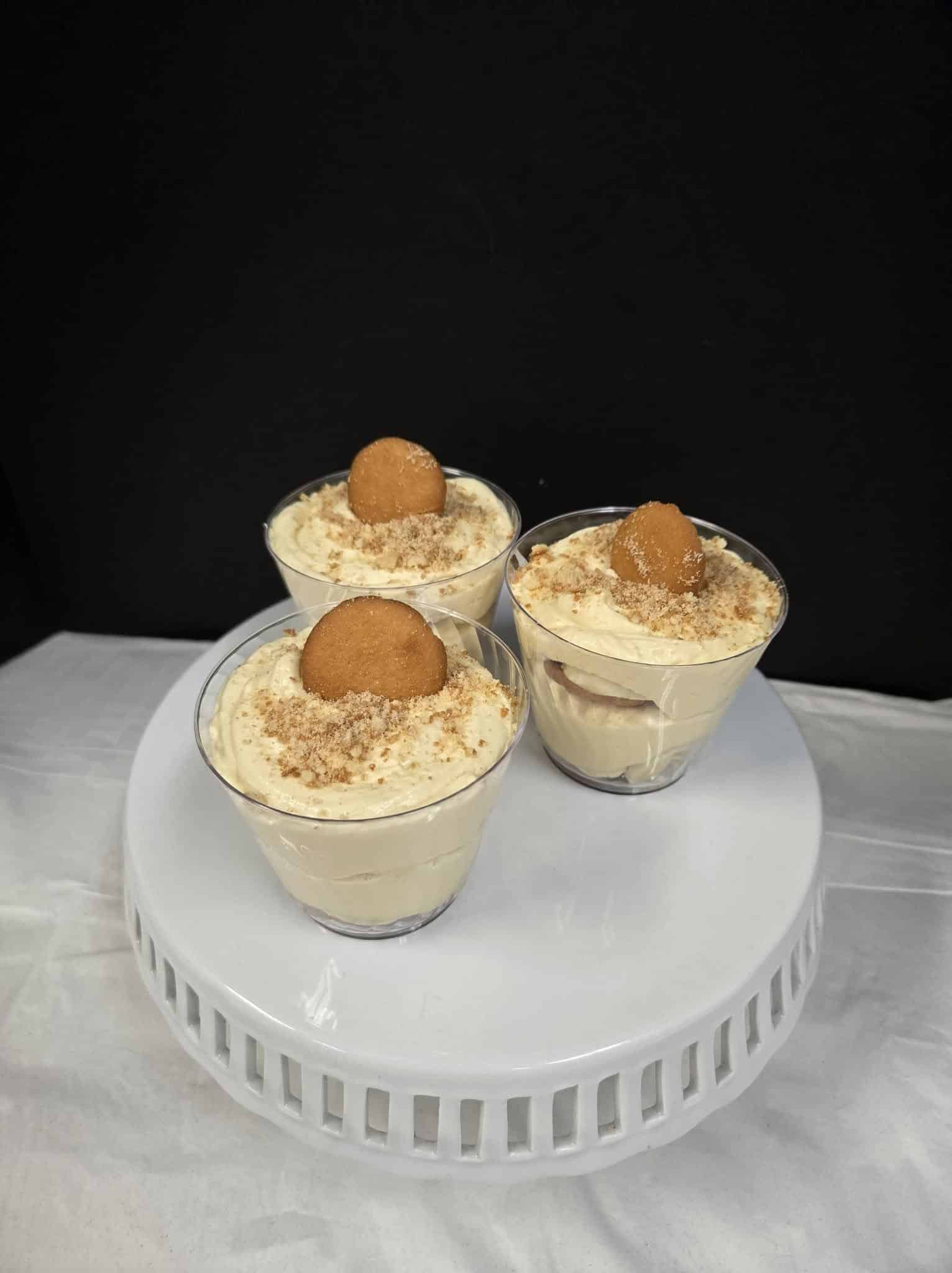 Three clear plastic cups of banana pudding on a stand, each garnished with vanilla wafers