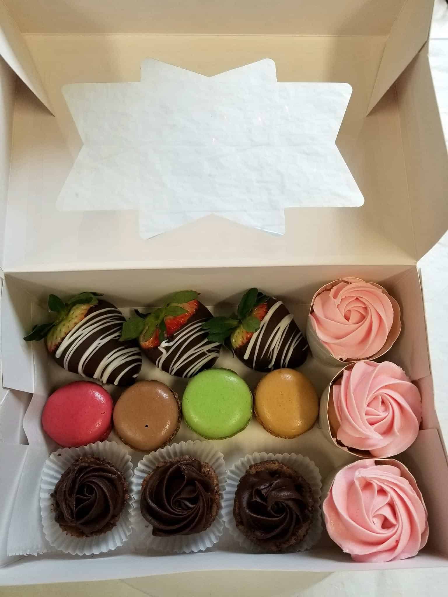 Bakery box of chocolate dipped strawberries, pink frosted cupcakes, colorful macarons, and chocolate frosted cupcakes