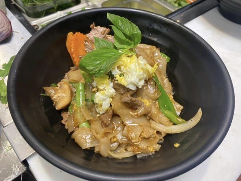 Black bowl of Thai noodles with pork, carrot, cabbage, and basil garnish in a brown sauce