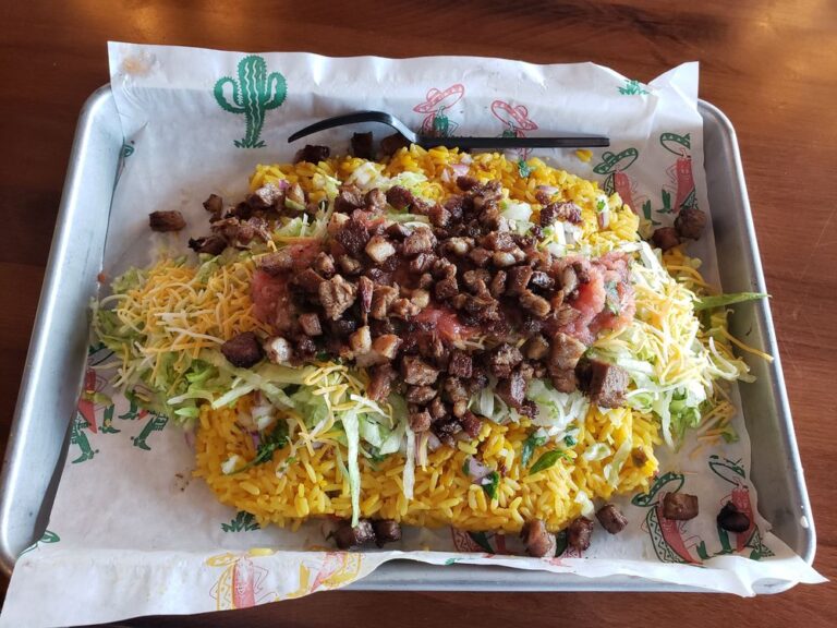 Mexican grilled pork bits on a bed of yellow rice with shredded lettuce and shredded mexican blend cheeses on branded parchment paper in a square stainless steel pan with a black plastic fork