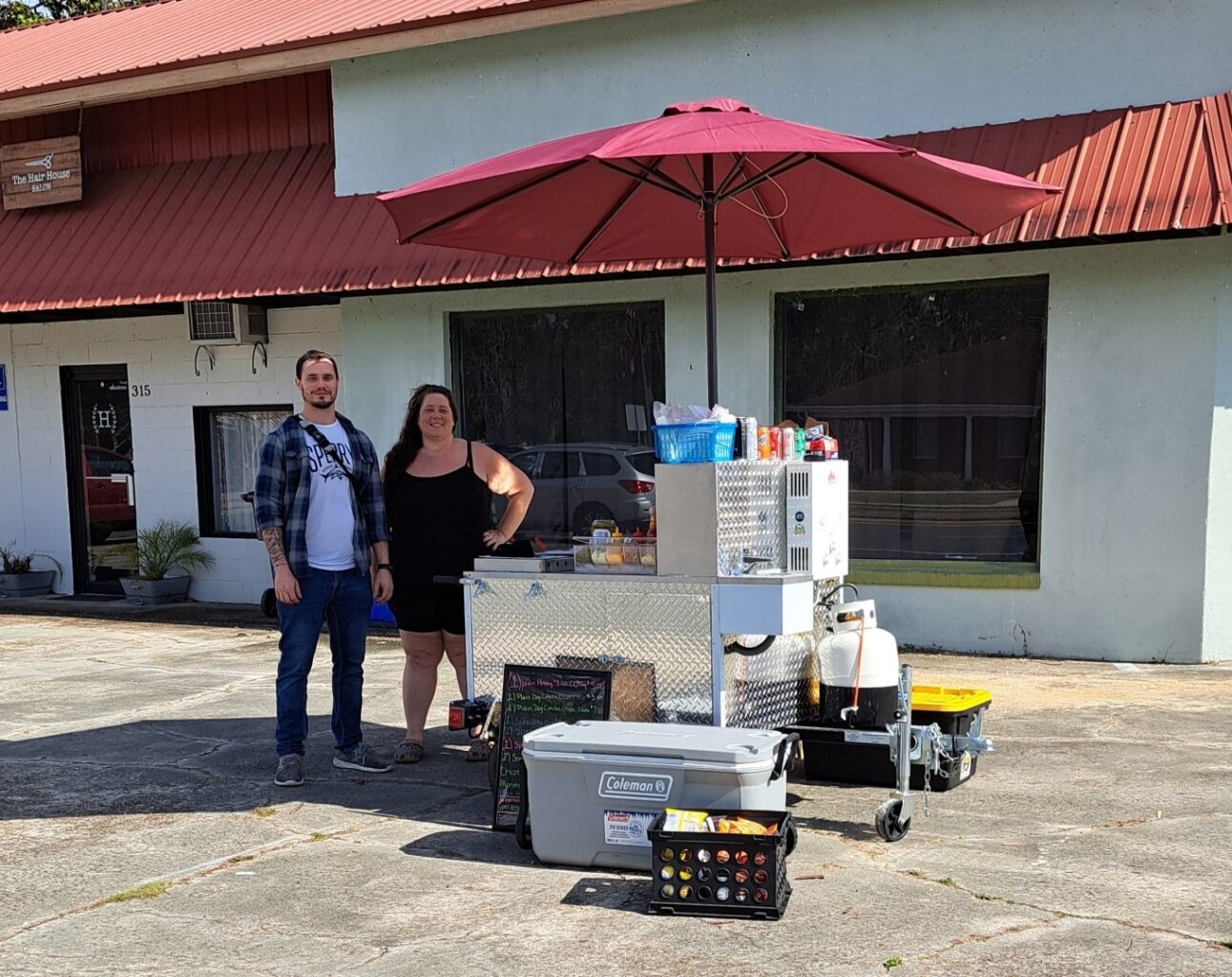 New Day Cafe outdoor food stand with red umbrella and two people standing by