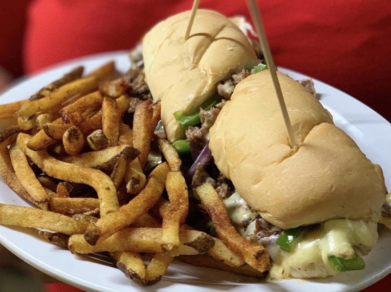 Horse and Cow Philly Cheese Steak Sub with green bell peppers and cheese on a white sub roll and sliced in half with toothpicks stuck in each half; also, French fries on the side, all on a white plate