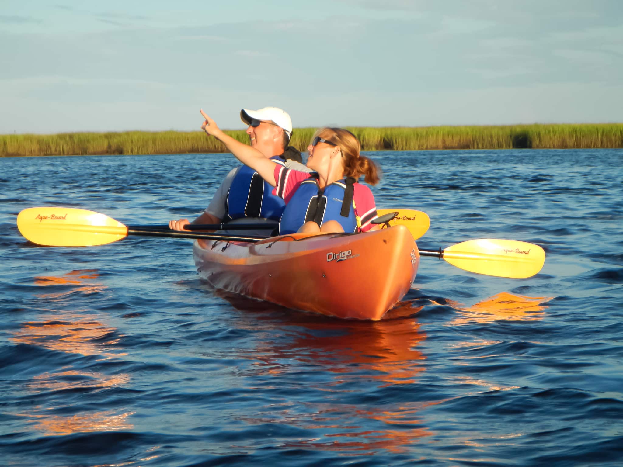 Man and woman in an orange kayak with yellow paddles. The woman is pointing to something interesting off the the viewer's left.