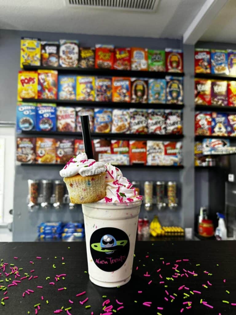 A milkshake in a clear plastic 16 oz Alien-Treats-branded cup, topped with whipped cream, sprinkles, and a cupcake garnish, sitting on a counter top with a candy display in the background