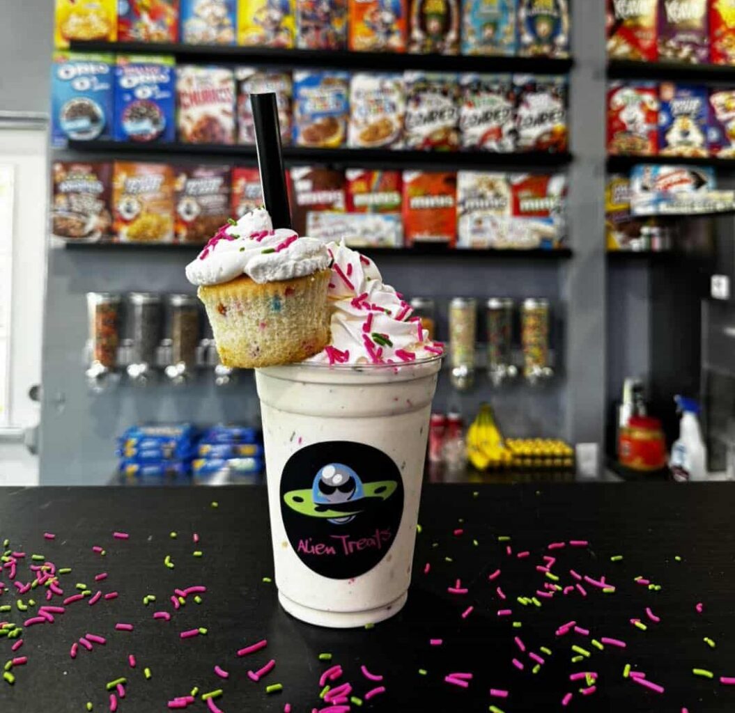 Milkshake with whipped cream and a cupcake garnish covered in sprinkles, and in a clear plastic Alien Treats-branded plastic cup with a black straw