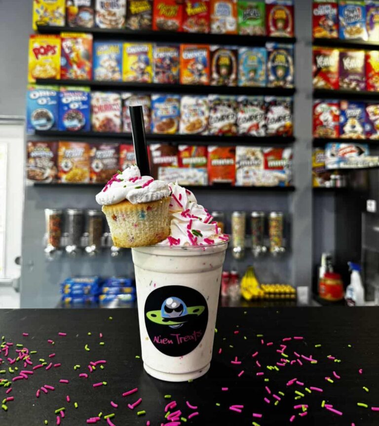 A milkshake in a clear plastic 16 oz Alien-Treats-branded cup, topped with whipped cream, sprinkles, and a cupcake garnish, sitting on a counter top with a candy display in the background