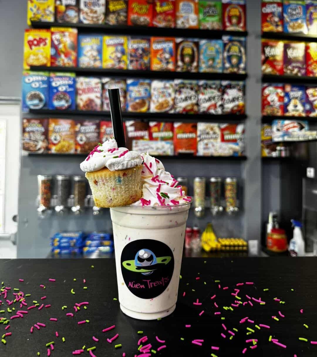 A milkshake in a clear plastic 16 oz Alien-Treats-branded cup, topped with whipped cream, sprinkles, and a cupcake garnish, sitting on a counter top with a candy display in the background