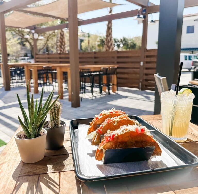 Three tacos on a tray, flanked by two cactus plants and a margarita on a wooden table outside at The Firefly Cantina