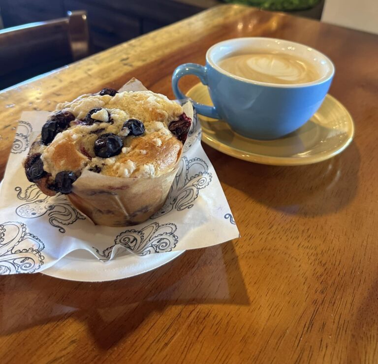Blueberry muffin on a paper and a plate and a latte in a blue coffee cup on a gold saucer all on a wood table