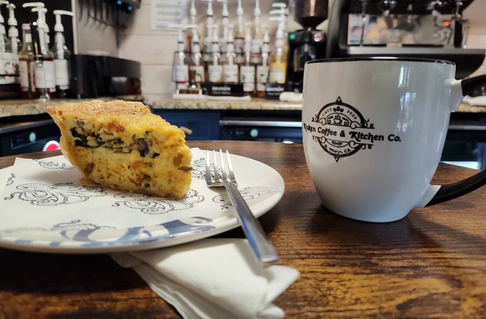 Branded Black and White Kraken Coffee Mug next to a slice of quiche on a plate with a fork