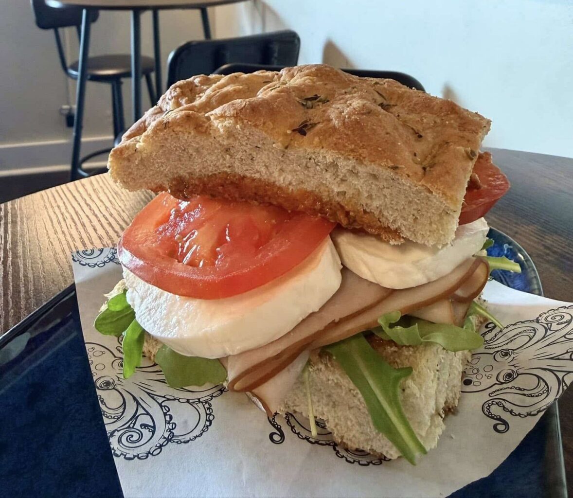 Turkey Mozzarella Sandwich with Aioli sauce and arugula on focaccia bread on Kraken Coffee-branded parchment paper on a black plate