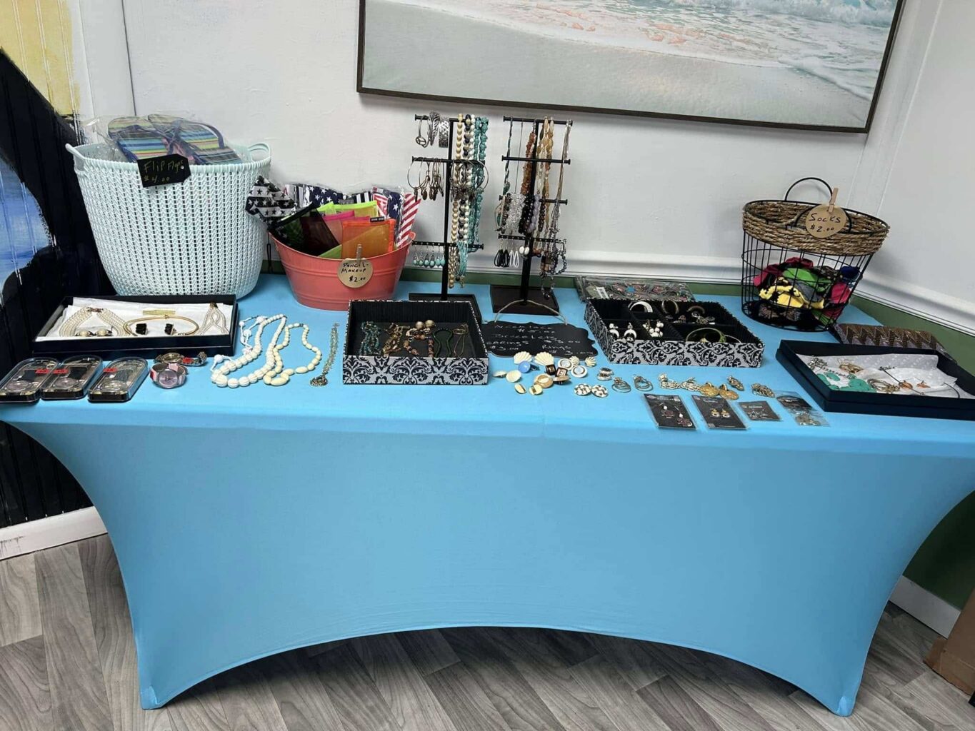 Display of jewelry and collectibles on a long table with a bright blue tablecloth in the corner of a room with white beadboard walls