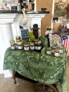 Display of jars of moonshine pickles and jellies in pint and quart jars, all sitting on a green table cloth decorated with ferns and other plant leaves