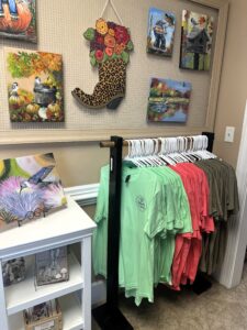 Rack of green and orange T-shirts in front of a wall hung with original paintings.