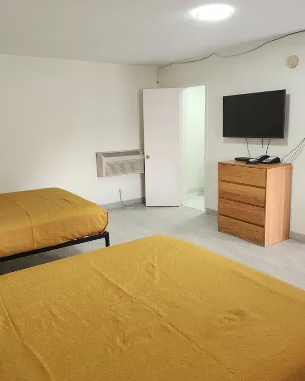 Economy Inn Double Room from inside showing two queen beds with gold colored blankets on simple black metal platform frames in the foreground left; and exterior door, wood dresser, flat screen tv at the far side of the room with white walls and a single overhead recessed LED ceiling light.