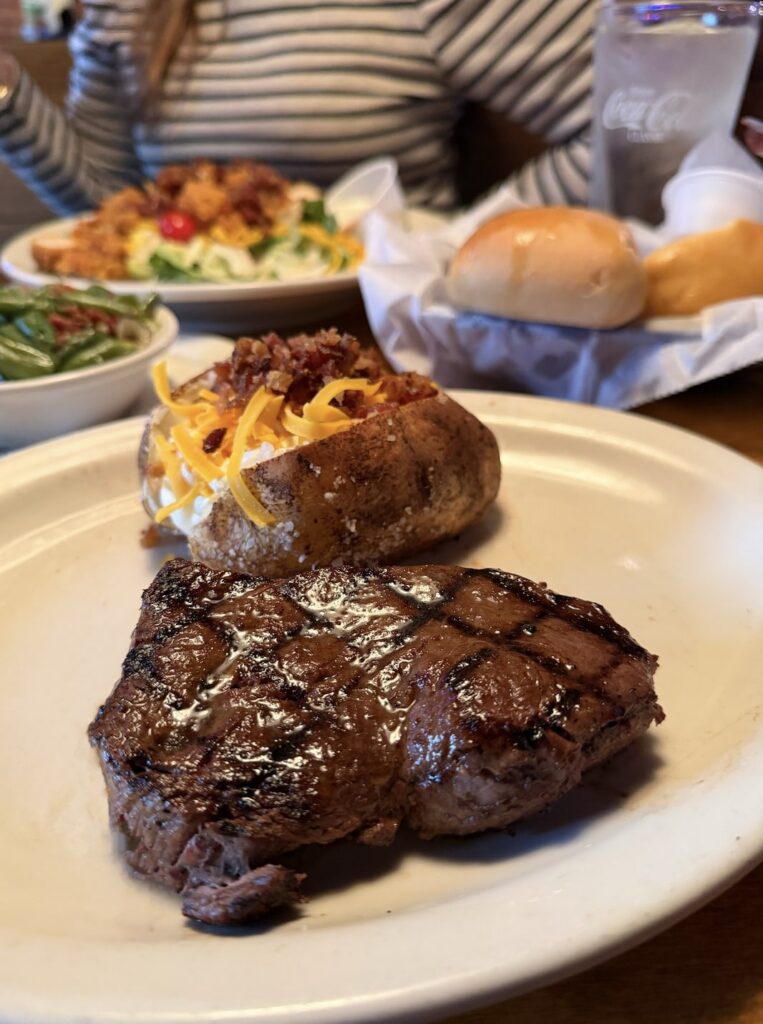 Texas Roadhouse steak with loaded baked potato. Rolls, salad and green beans are in the back ground.