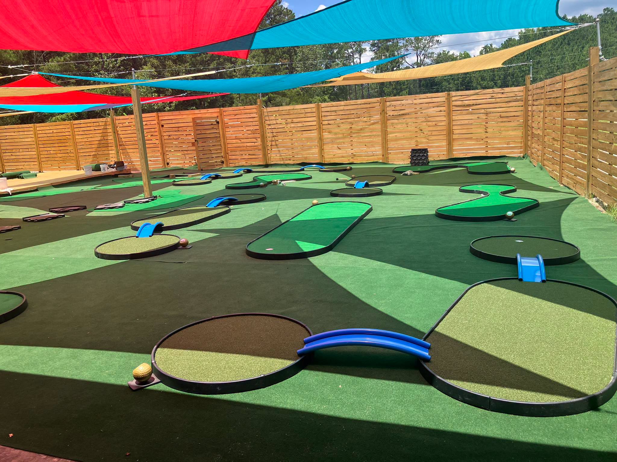 Outdoor mini golf course surrounded by a wooden fence and shaded by red, blue, and yellow sails overhead.