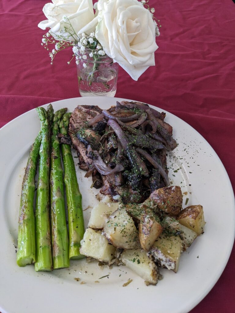 T-bone steak topped with sauteed vegies on a white plate with roasted potatoes and asparagus spears