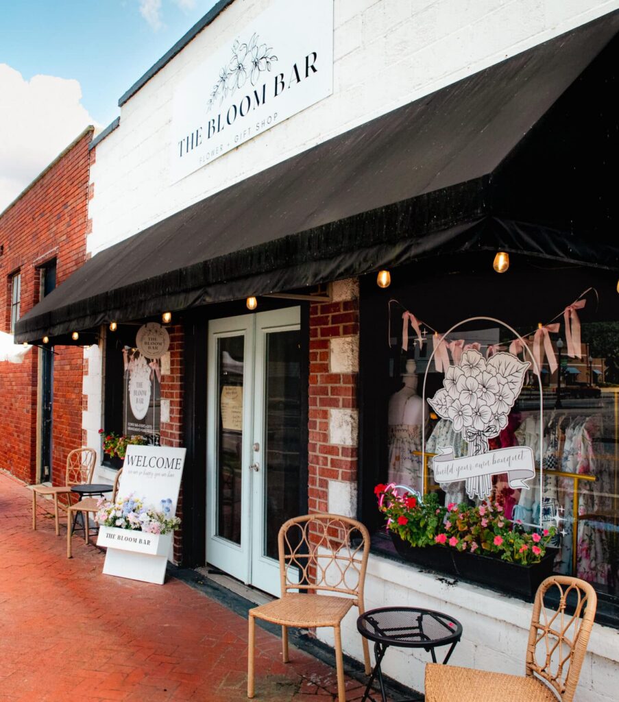 Exterior shot of florist shop store front with black awning and red brick sidewalk and a white patio umbrella.