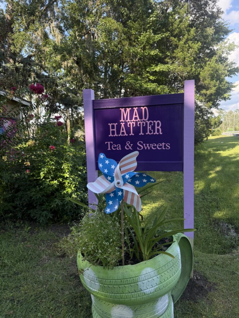Purple and pink outdoor sign for Mad Hatter Tea and Sweets Boutique with large green planter and a pinwheel decorated with stars and stripes