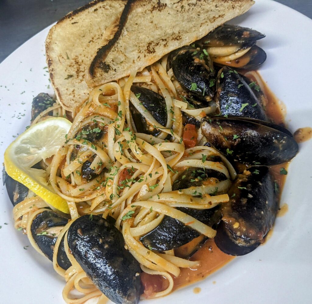 Shallow white bowl of pasta el diable with mussels in a red sauce with two pita triangles and a lemon wedge garnish at Seagles Restaurant