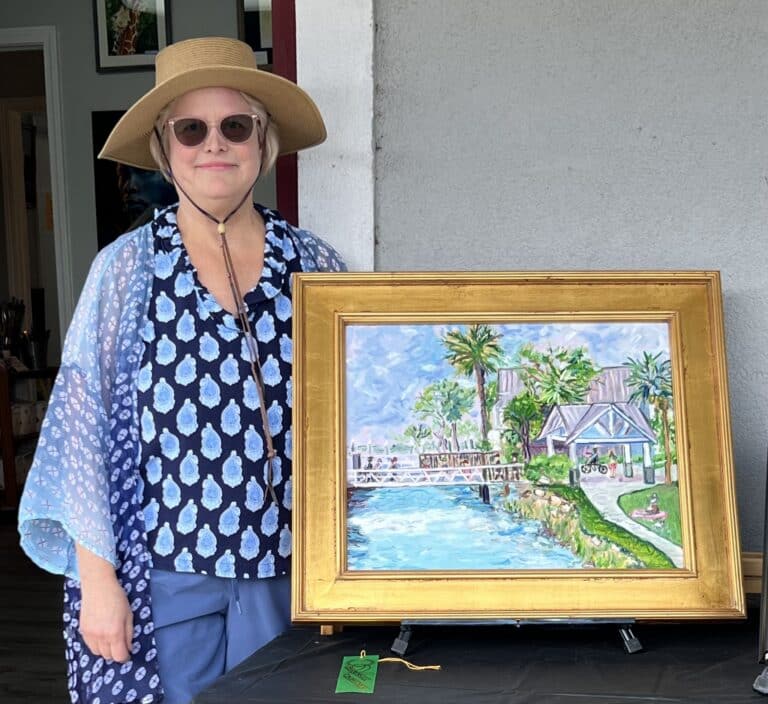Artist Jennie Bagwell dresy in blue, with a straw hat and sunglasses holding one of her framed paintings