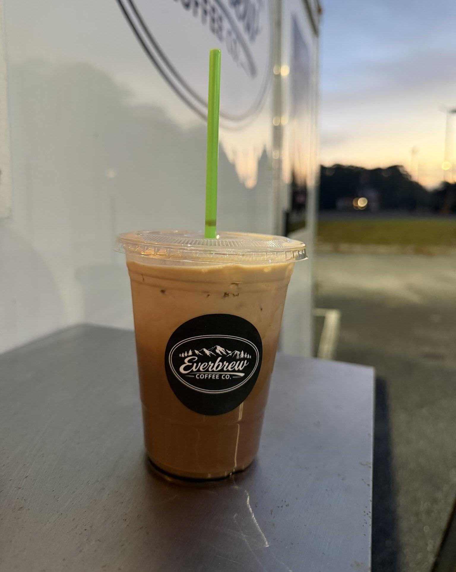 Chunky Monkey iced coffee in a clear plastic cup on the exterior counter of a food truck with the sunrise in the background.